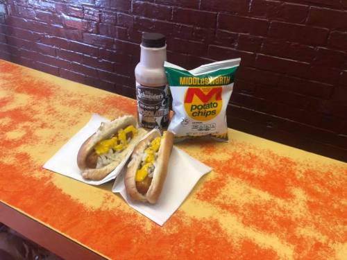 Two hot dogs with toppings, a bottle of mustard, and a bag of potato chips on a colorful table.
