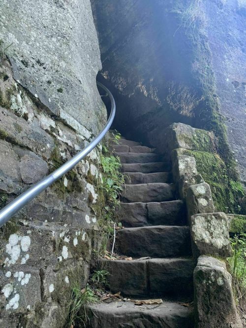 Stone steps lead into a rocky crevice, with a metal handrail and greenery surrounding the entrance.