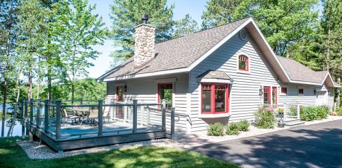 A charming house with a stone chimney, red accents, and a deck surrounded by trees near a calm lake.