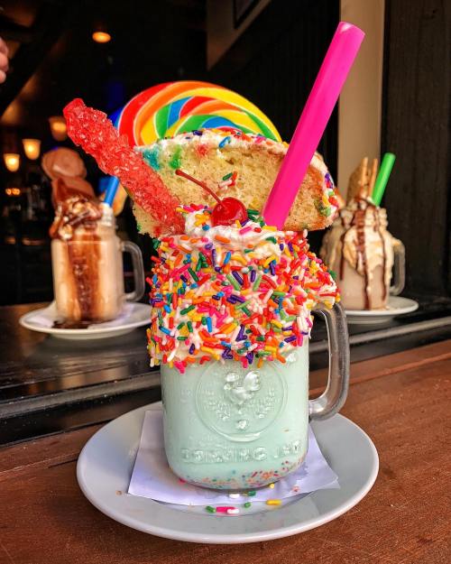 A colorful milkshake topped with whipped cream, sprinkles, a cherry, a cookie, and a rainbow lollipop in a glass jar.
