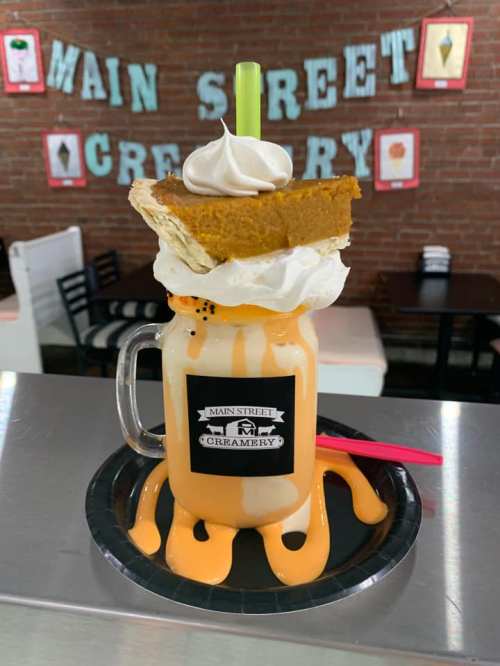 A colorful milkshake topped with whipped cream, a slice of pie, and drizzled with orange sauce, served in a mason jar.