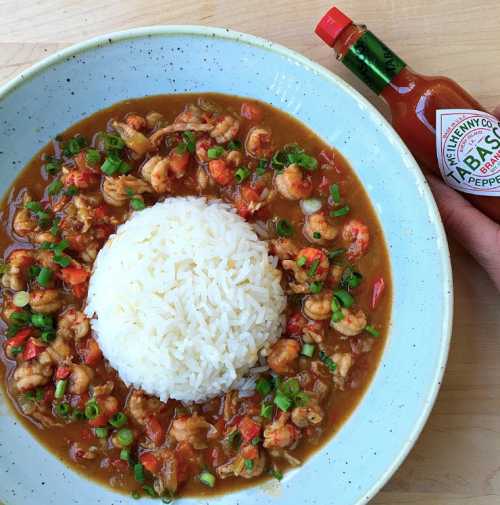 A bowl of shrimp stew with rice, garnished with green onions, alongside a bottle of hot sauce.