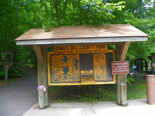 Sign at Timm's Hill County Park displaying maps and information about the park, surrounded by lush greenery.
