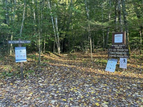 Trail entrance to a waterfall, with signs indicating prohibited activities like camping and fires, surrounded by trees.