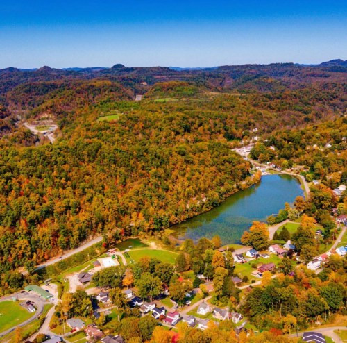 Aerial view of a serene lake surrounded by colorful autumn foliage and quaint houses in a hilly landscape.