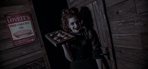 A woman with curly hair holds a tray of pastries in a dimly lit, rustic setting, with a sign for meat pies in the background.