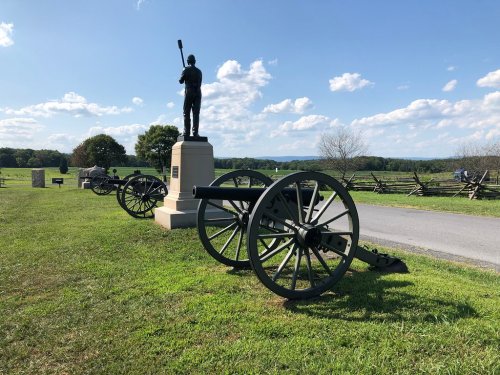 A soldier statue stands beside historic cannons on a grassy field under a blue sky with scattered clouds.