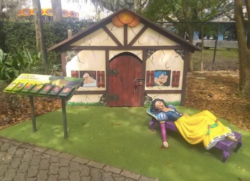 A whimsical scene featuring a painted cottage and a statue of Snow White resting on a bench in a garden setting.