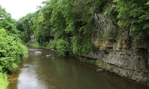 A serene river flows between lush green trees and rocky cliffs, creating a peaceful natural landscape.