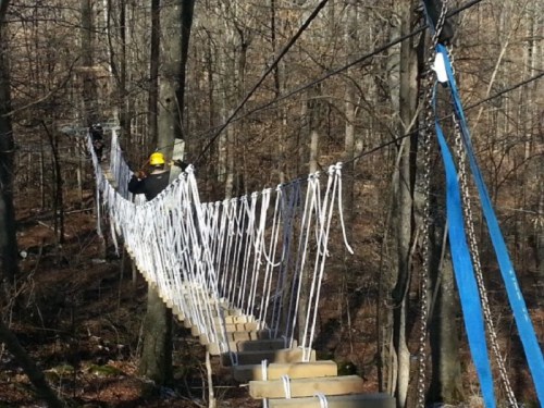 A person in a hard hat walks across a suspended rope bridge in a wooded area.