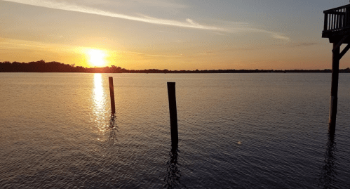 Sunset over a calm lake, with silhouetted posts in the water and a pier in the foreground.