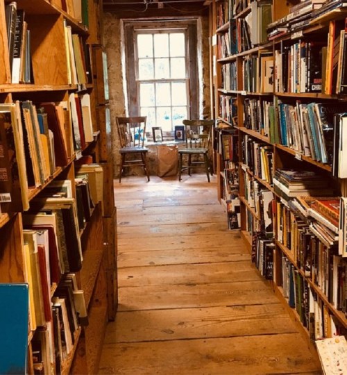 A cozy bookstore aisle lined with wooden shelves filled with books, leading to a window and two chairs.