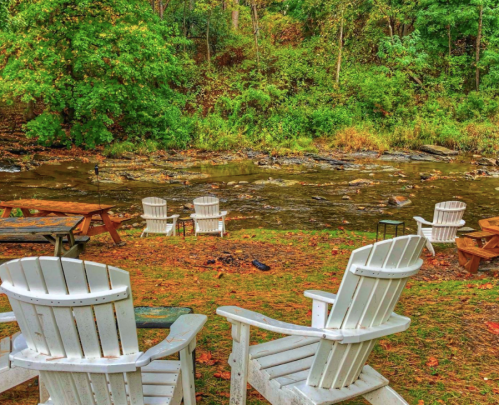 A serene riverside scene with white Adirondack chairs and lush greenery in the background.
