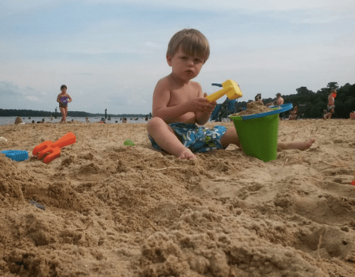 A young boy sits on the beach, playing with a bucket and toys in the sand, while others enjoy the water in the background.