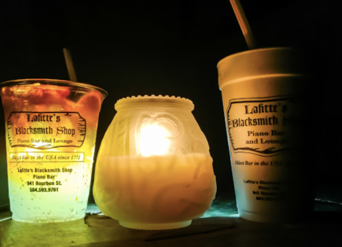 A glowing candle beside two drinks, one in a clear cup and the other in a white cup, set against a dark background.