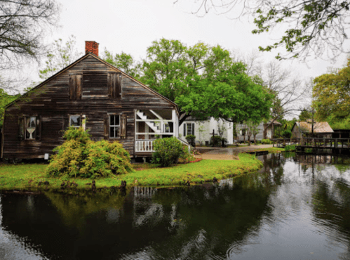 A rustic wooden house beside a calm pond, surrounded by lush greenery and trees on a cloudy day.