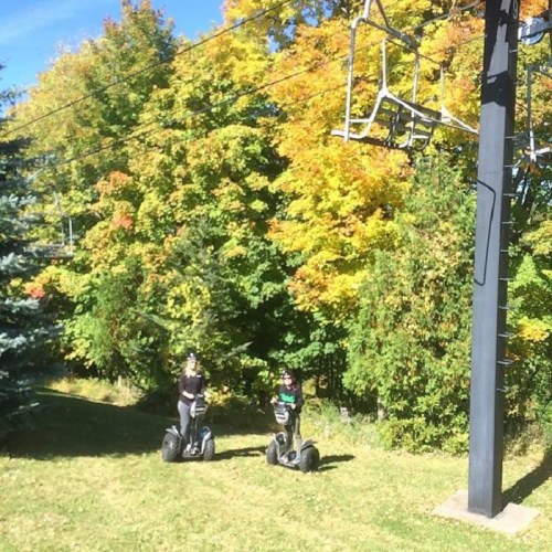 Two people on segways in a grassy area with colorful autumn trees and a ski lift in the background.