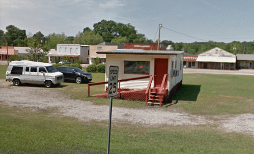 A small, red and white building with a porch, surrounded by grass and parked cars, near a speed limit sign.