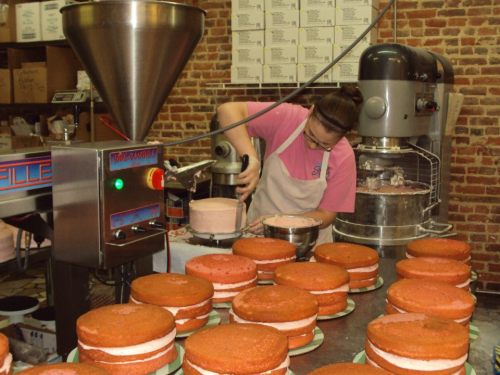 A baker decorates layered cakes with frosting in a bakery, surrounded by finished cakes on a table.