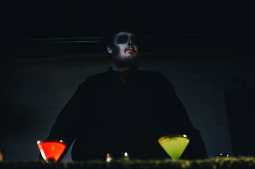 A person with skull makeup stands behind a table with colorful cocktails, illuminated in dim lighting.