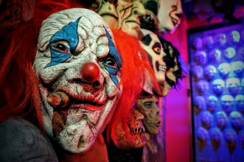 A close-up of a creepy clown mask with orange hair, surrounded by various eerie masks in a dimly lit setting.