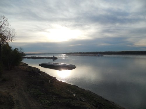 A serene river scene at sunset, with a barge on the water and soft clouds reflecting in the calm surface.
