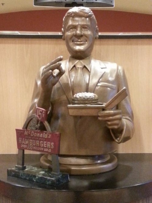 A bronze statue of a smiling man holding a hamburger and a tray, with a sign reading "McDonald's Hamburgers."