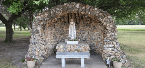 A stone grotto with a statue of Mary, surrounded by flowers and trees in a grassy area.