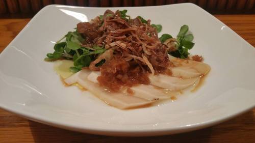 A white bowl with sliced chicken, topped with crispy shallots and a savory sauce, served on a bed of greens.