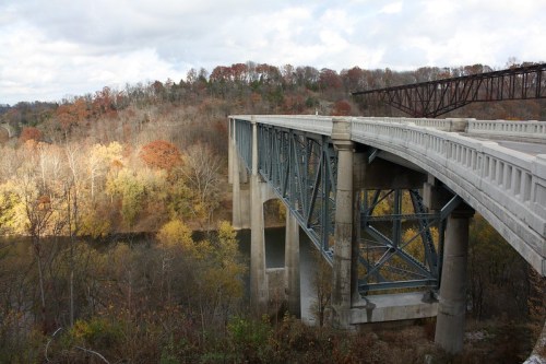 A modern bridge spans a river, surrounded by autumn foliage and rolling hills under a cloudy sky.