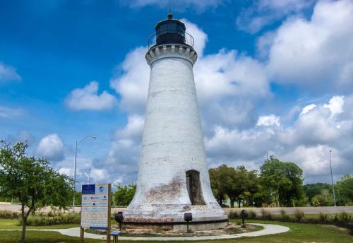A tall white lighthouse stands against a blue sky with clouds, surrounded by greenery and a sign nearby.