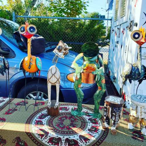 Colorful metal sculptures of a bird, frog, and other whimsical characters displayed on a table, with a blue van in the background.