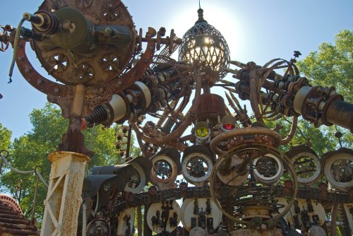 A large, intricate sculpture made of metal gears and pipes, illuminated by sunlight from behind.