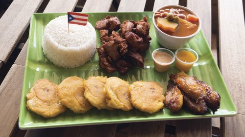 A vibrant plate of Puerto Rican food featuring rice, fried pork, tostones, and a bowl of bean stew, garnished with a flag.