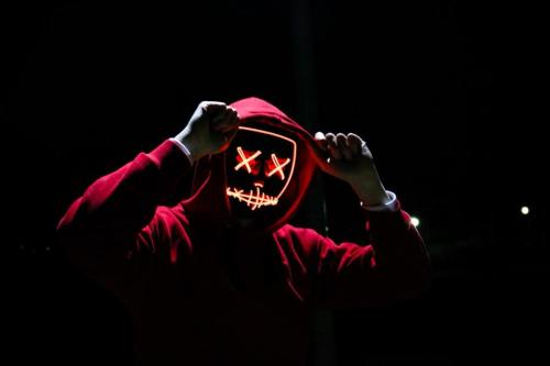 A person in a red hoodie poses at night, wearing a glowing mask with a sinister smile and crossed-out eyes.