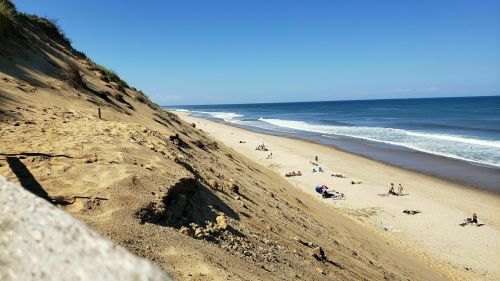 A sandy beach with gentle waves, people sunbathing, and a steep dune in the background under a clear blue sky.