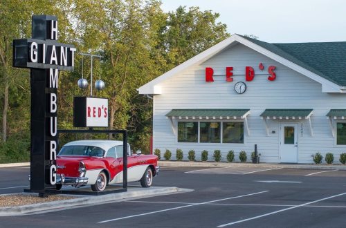A vintage red car parked outside a diner named "Red's," featuring a large sign that reads "GIANT HAMBURG."