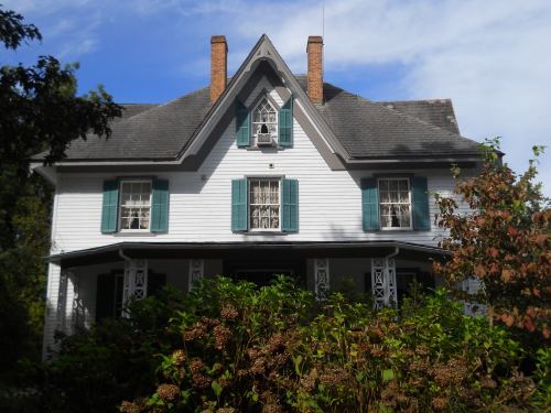A charming white house with green shutters, a steep roof, and surrounded by lush greenery and flowers.