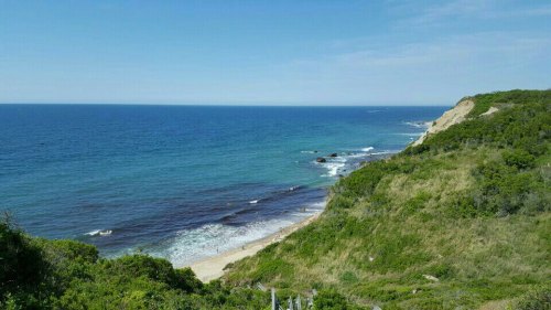 A scenic view of a coastline with blue ocean, sandy beach, and lush green cliffs under a clear sky.