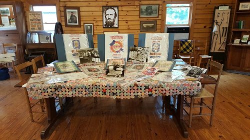 A wooden table displays various historical documents, photos, and memorabilia in a rustic room with wooden walls.