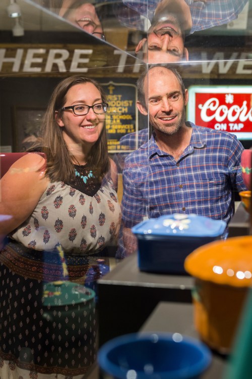 A man and woman smile while looking at colorful pottery displayed in a glass case, with reflections visible.