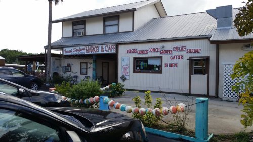 A quaint market and café with a sign displaying various seafood options, surrounded by palm trees and parked cars.