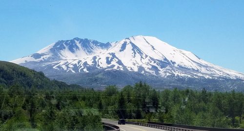 Snow-capped mountains rise above a green landscape under a clear blue sky, with a winding road in the foreground.