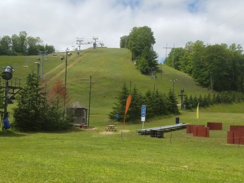A grassy ski slope with chairlifts, surrounded by trees and a picnic area, under a cloudy sky.