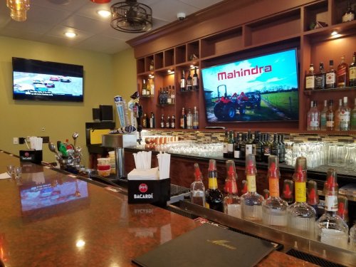 A bar with a polished countertop, shelves of liquor, and two screens displaying a tractor ad and a racing event.