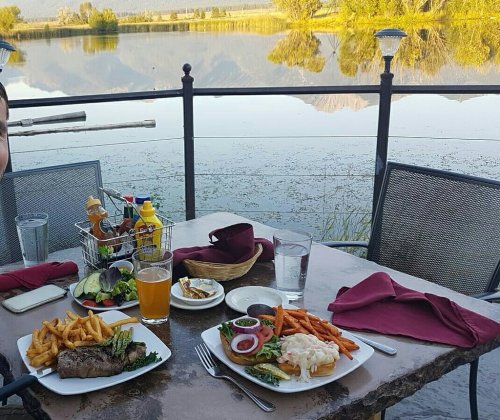 A lakeside dining table with steak, sweet potato fries, salad, and drinks, surrounded by scenic nature.
