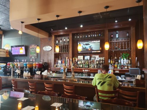 A bar with a wooden counter, shelves of liquor, and a TV showing a race, with a person sitting at the bar.