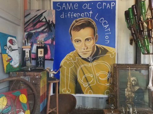 A colorful art display featuring a large portrait with the text "Same ol' crap, different location" and various artworks.