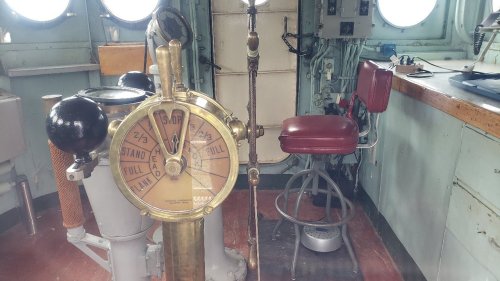 A vintage ship's wheel with a brass indicator and a red stool in a nautical control room.
