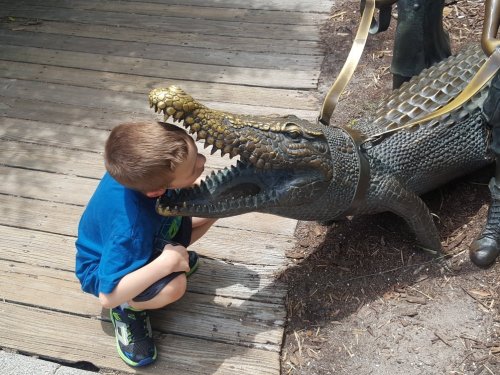 A young boy leans in to kiss a bronze alligator statue on a wooden walkway.